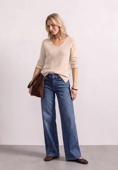 Femme souriante en pull beige à col V, jean large et sac marron sur fond clair.