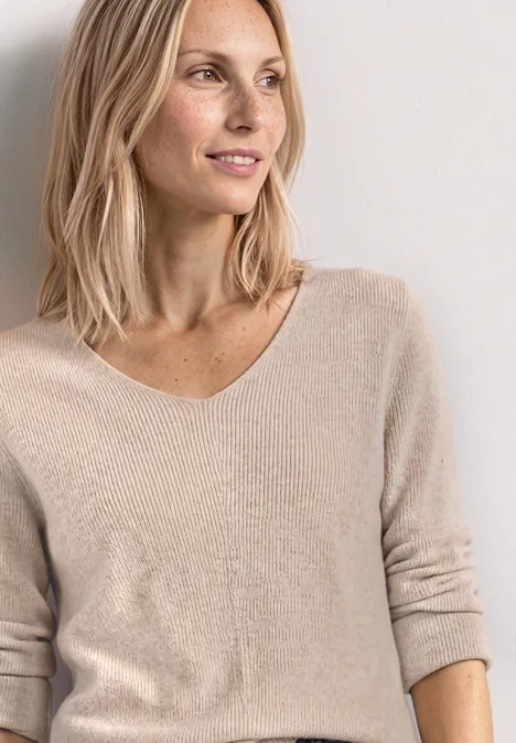 Femme blonde élancée, portant un pull beige à col V, regardant légèrement sur le côté avec un sourire.