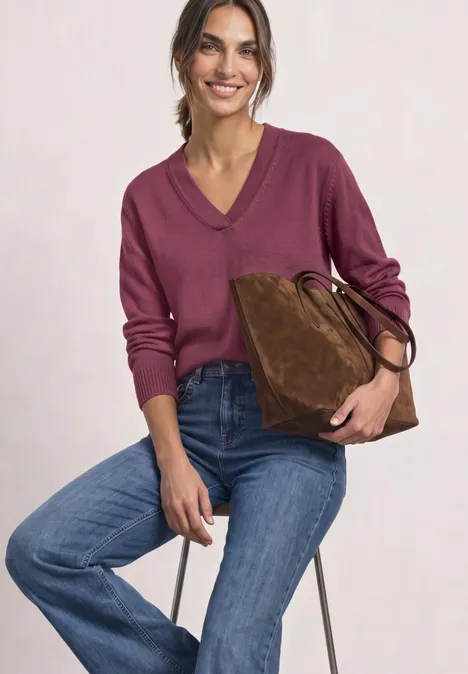 Langarm Pullover mit V-Ausschnitt dark clouded blush Frau sitzt auf Hocker, trägt bordeauxroten Pullover, blaue Jeans und hält eine braune Wildledertasche.