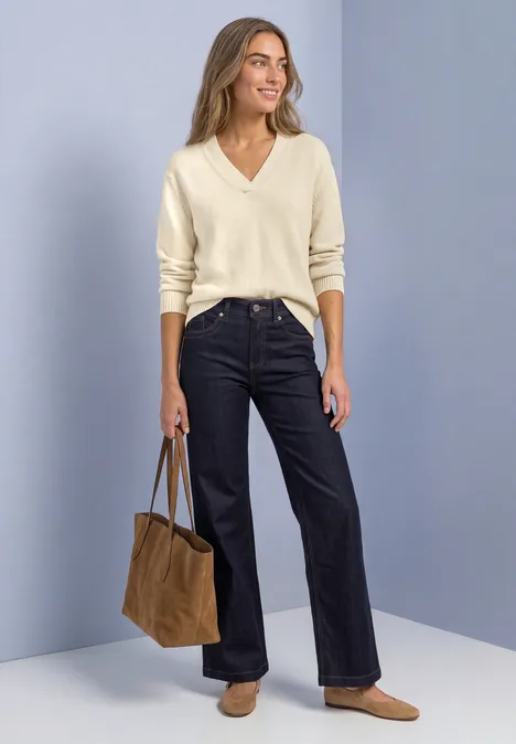 Frau in cremefarbenem V-Pullover, dunklen weiten Jeans, beigen Slippern und brauner Tasche vor schlichtem Hintergrund.
