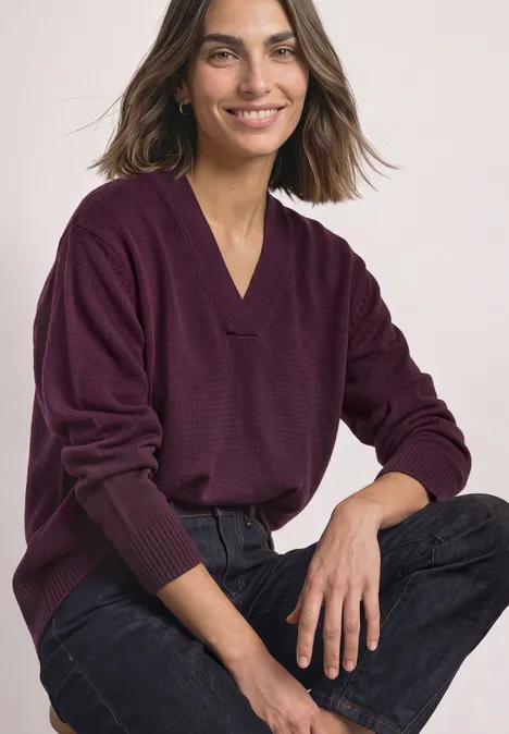 Eine lächelnde Frau im violetten V-Pullover und schwarzen Jeans sitzt entspannt.