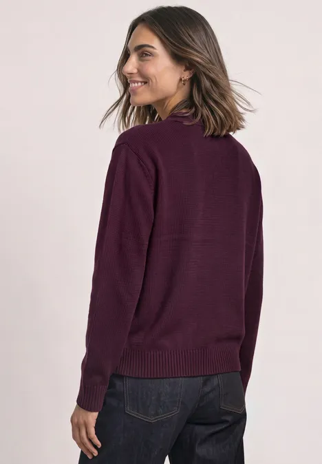 Frau mit schulterlangem Haar trägt einen violetten Pullover und dunkle Jeans, sie lächelt über die Schulter.