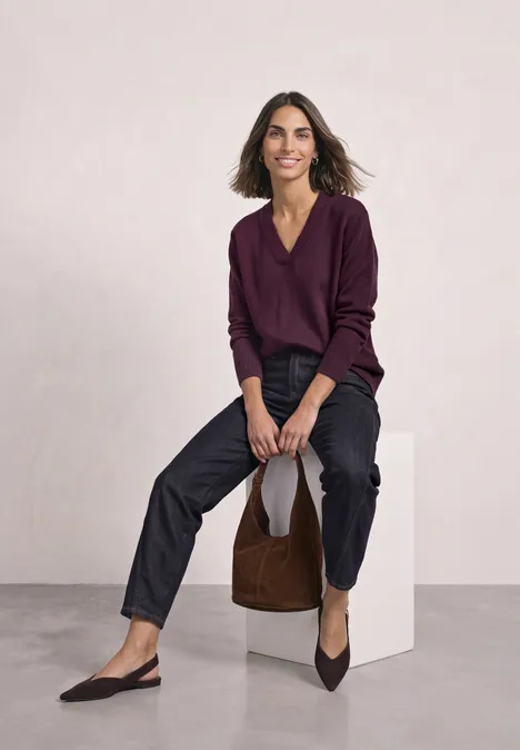 Frau sitzt auf weißem Würfel, trägt violetten Pulli, schwarze Hose, spitze Ballerinas und elegante Tasche.