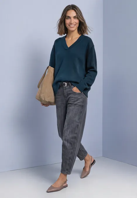 Frau in lässigem Outfit mit dunkelblauem V-Pullover, grauen Hosen, beigen Ballerinas und Tasche vor hellem Hintergrund.