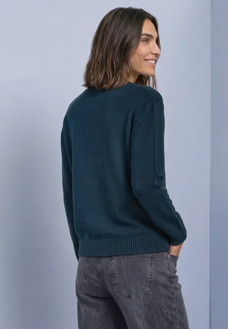 Frau von hinten, trägt einen dunkelblauen Pullover und graue Jeans vor hellem Hintergrund.
