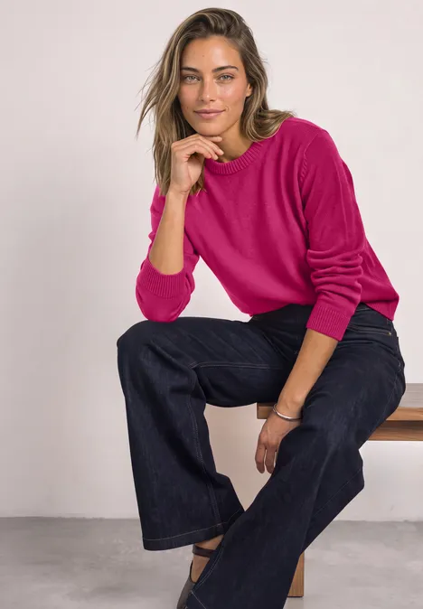 Frau in pinkem Pullover und weiten Jeans sitzt lässig auf einer Bank vor hellem Hintergrund.