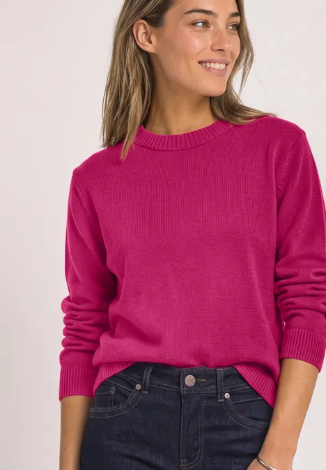 Eine lächelnde Frau trägt einen pinken Pullover und dunkelblaue Jeans.