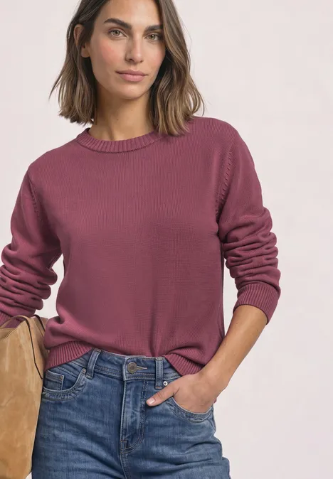 Frau in rosa Pullover und blauen Jeans lehnt lässig, eine Hand in der Tasche.