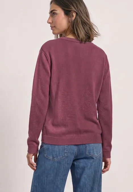Frau von hinten mit kurzem dunklem Haar trägt violetten Pullover und blaue Jeans vor hellem Hintergrund.