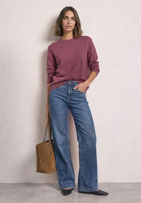 Frau steht vor weißer Wand, trägt bordeauxfarbenen Pullover, weite Jeans und hält eine braune Tasche.