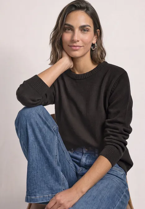 Frau mit braunem Pullover und Jeans sitzt lässig und blickt in die Kamera.