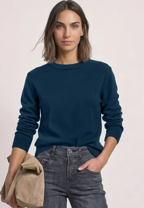 Langarm Pullover mit Boat Neck blue petrol Frau in marineblauem Pullover und grauer Jeans, hält eine beige Jacke, vor hellem Hintergrund.