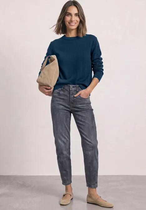 Langarm Pullover mit Boat Neck blue petrol Frau in dunkelblauem Pullover, grauen Jeans und beigen Slippern hält eine beige Tasche vor neutralem Hintergrund.