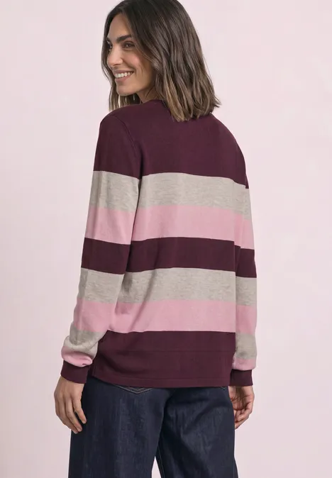 Eine Frau trägt einen breit gestreiften Pullover in Bordeaux, Rosa und Beige, schaut lächelnd über die Schulter.