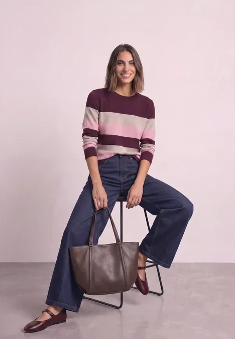 Frau in gestreiftem Pullover, weiten Jeans und Slippern sitzt lächelnd auf Hocker, hält braune Ledertasche.