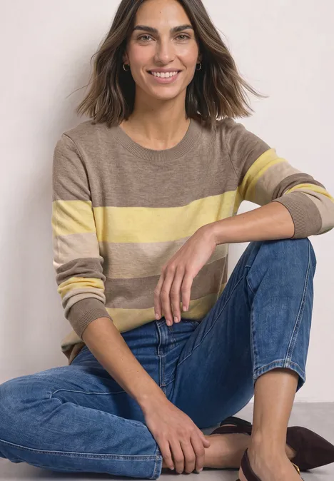 Eine lächelnde Frau in gestreiftem Pullover und Jeans sitzt entspannt auf dem Boden.