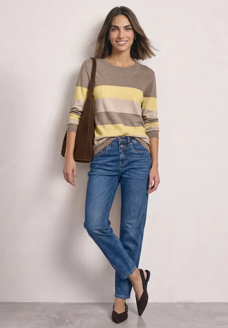 Frau in gestreiftem gelb-beige Pullover, blauen Jeans und schwarzen Schuhen vor hellem Hintergrund.