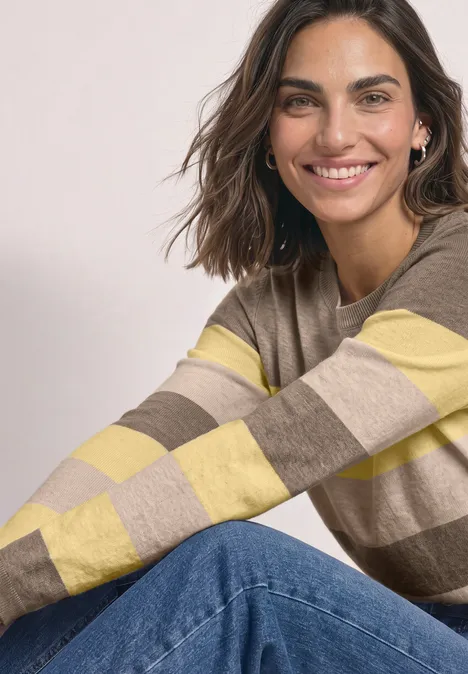 Eine lächelnde Frau trägt einen gestreiften beige-gelben Pullover und Jeans und sitzt entspannt.