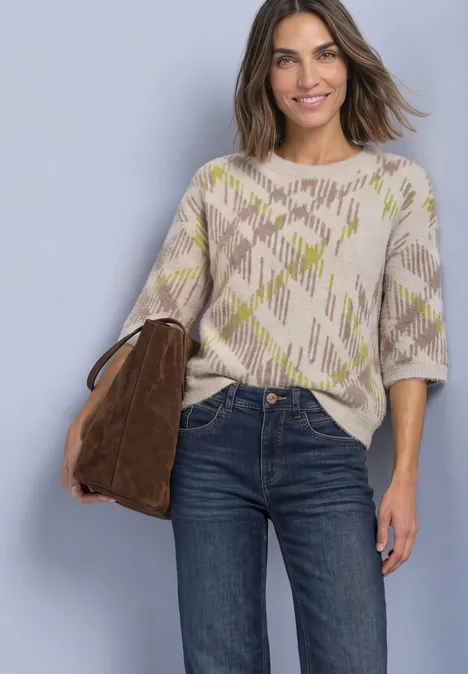 Frau in Beige-Grau gemustertem Pullover, dunklen Jeans und brauner Tasche lächelt vor hellgrauem Hintergrund.