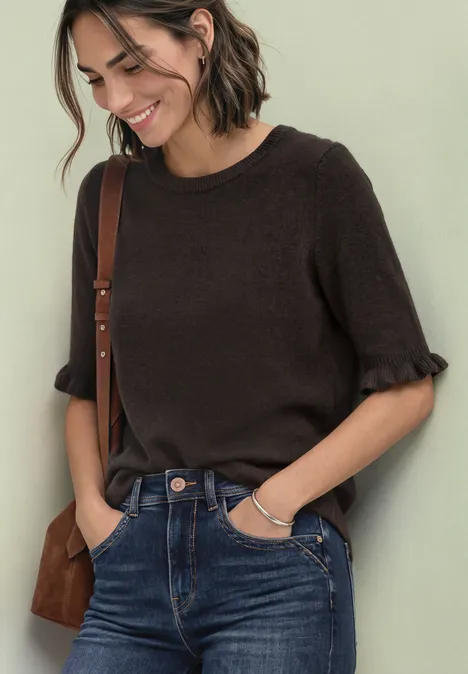 Vrouw in bruin shirt met ruches, spijkerbroek en tas, glimlachend voor een lichte achtergrond.