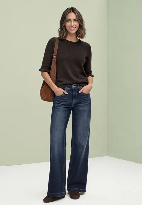 Vrouw met bruin kort mouwtricot, wijde jeans en bruine schoudertas voor een lichte achtergrond.