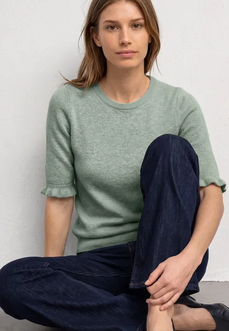 Kurzarm Pullover mit Rundhals und Rüschen eucalyptus green mel. Frau in grünem Kurzarm-Pullover und blauen Jeans sitzt entspannt mit verschränkten Beinen vor weißem Hintergrund.