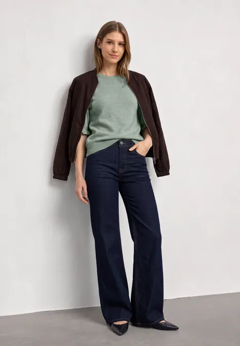 Kurzarm Pullover mit Rundhals und Rüschen eucalyptus green mel. Eine Frau steht vor weißer Wand, trägt grünen Pullover, Jacke über den Schultern und weite Jeans.