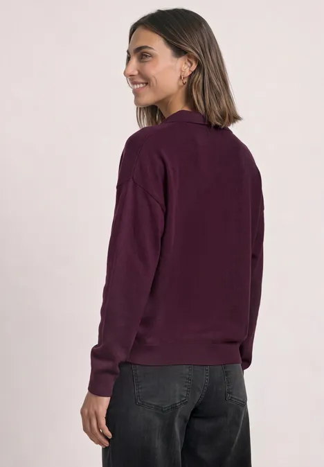 Jonge vrouw in zijaanzicht draagt een bordeauxrood shirt met lange mouwen en een donkere broek.