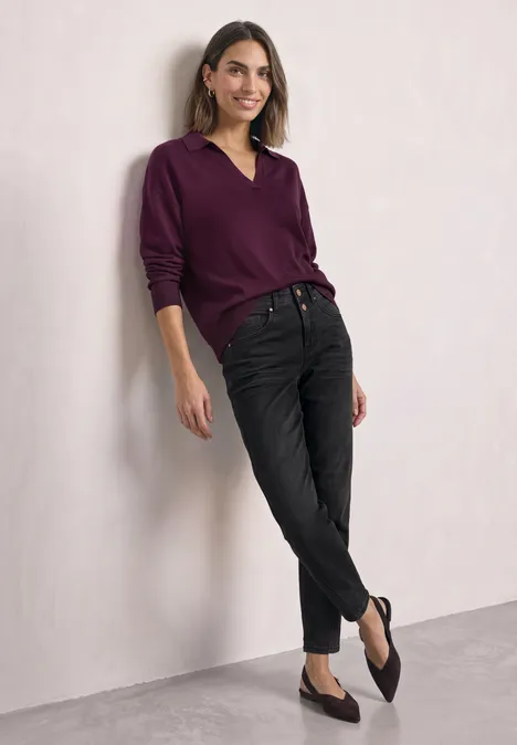 Een glimlachende vrouw in een paarse trui leunt ontspannen tegen een muur, draagt zwarte jeans en platte schoenen.