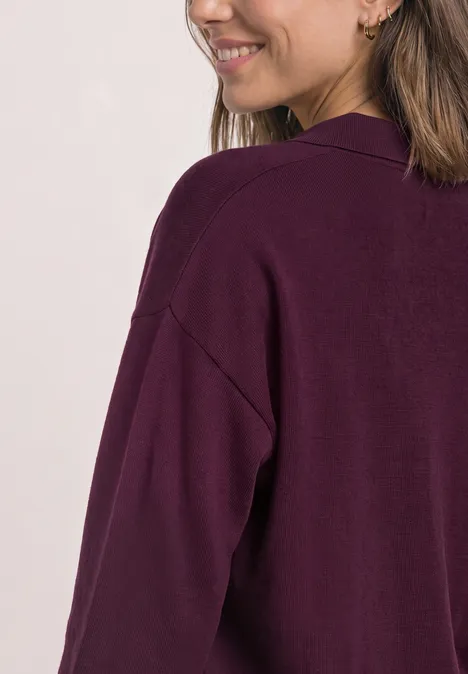 Vrouw van opzij/achterkant in een losvallende donkerpaarse cardigan, glimlachend, schouders zichtbaar.