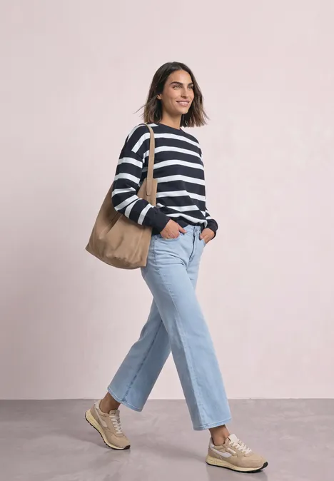 Vrouw in gestreepte trui, jeans, sneakers en shopper, glimlachend lopend.