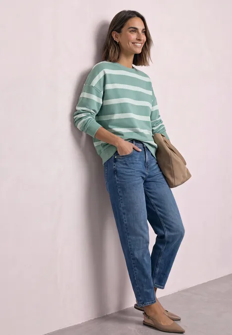 Sweatshirt mit Rundhals und Streifen eucalyptus green Frau lehnt lächelnd an Wand, trägt grünen Ringelpullover, blaue Jeans und braune Slipper.