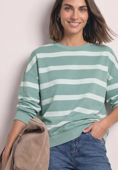 Sweatshirt mit Rundhals und Streifen eucalyptus green Frau trägt grünen weiß gestreiften Pullover, blaue Jeans, lächelt, hält eine beigefarbene Tasche.