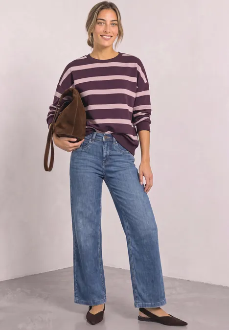 Bluza z okrągłym dekoltem i w paski berry maroon Kobieta w fioletowym swetrze w paski, szerokich niebieskich dżinsach i czarnych butach, z brązową torbą na jasnym tle.