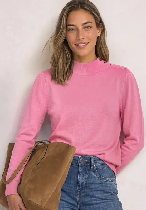 Een glimlachende vrouw draagt een roze trui, jeans en heeft een bruine tas over haar schouder.