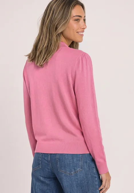 Vrouw in profiel met roze trui en blauwe spijkerbroek tegen lichte achtergrond.