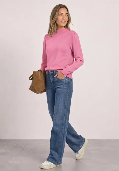 Jonge vrouw draagt roze trui, blauwe jeans en witte sneakers, met een bruine tas.