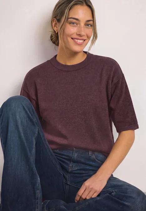 Pull à manches 3/4 et col rond rustic rouge mel. Junge Frau sitzt lässig, trägt lila Pullover und blaue Jeans, lächelt entspannt in die Kamera.
