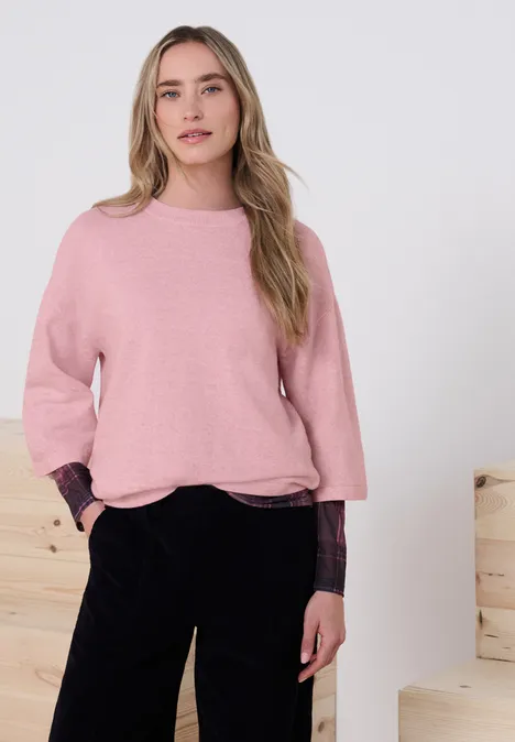 Junge Frau im rosa Pullover und schwarzen Hosen steht lässig mit Händen in den Taschen vor hellen Holzblöcken.