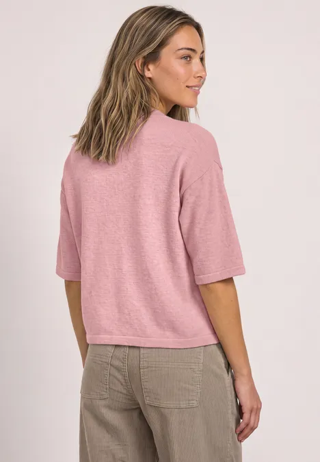 Eine Frau im rosa T-Shirt und beigen Hosen steht leicht seitlich und schaut zur Seite.