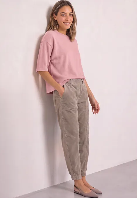 Eine Frau lehnt lächelnd an einer Wand, trägt ein rosa Oberteil, graue Cordhosen und graue Ballerinas.