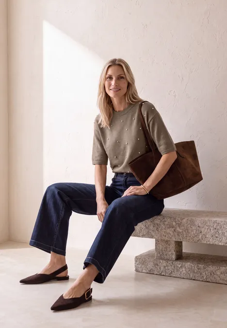 3/4-Arm Pullover mit Rundhals sanded amber mel. Blonde Frau in grünem Pullover, Jeans und brauner Tasche sitzt auf einer Bank in hellem Raum.