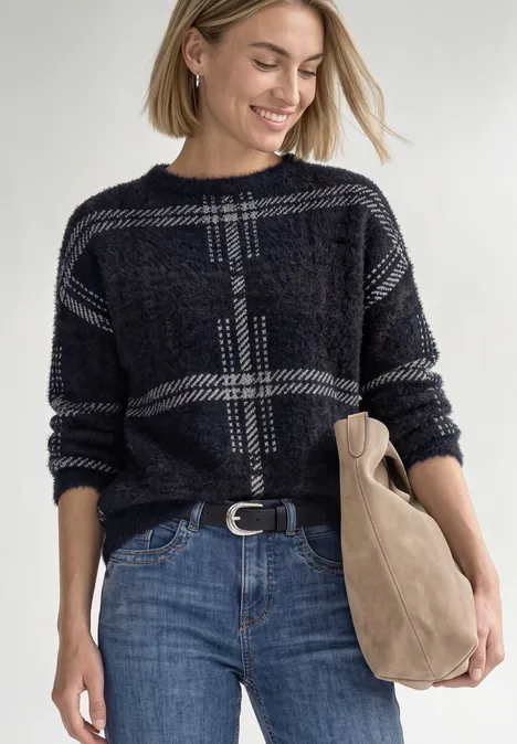 Een lachende vrouw met blond haar draagt een geruit trui, blauwe jeans en houdt een tas vast.