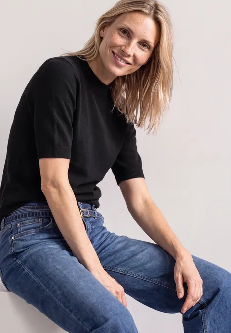 Pull à manches courtes et col roulé Black Eine blonde Frau in schwarzem T-Shirt und Jeans sitzt lächelnd vor hellem Hintergrund.