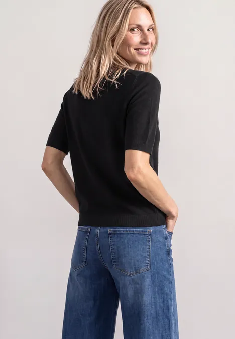 Pull à manches courtes et col roulé Black Eine Frau steht mit dem Rücken zur Kamera, schaut über die Schulter, trägt ein schwarzes Shirt und blaue Jeans.