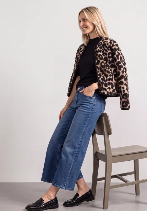 Pull à manches courtes et col roulé Black Frau lehnt lässig an einem Hocker, trägt Leo-Jacke, schwarzen Pullover, weite Jeans und schwarze Schuhe.