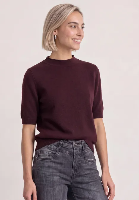 Eine junge Frau mit kurzem blondem Haar trägt einen dunklen Pullover und ausgewaschene Jeans.