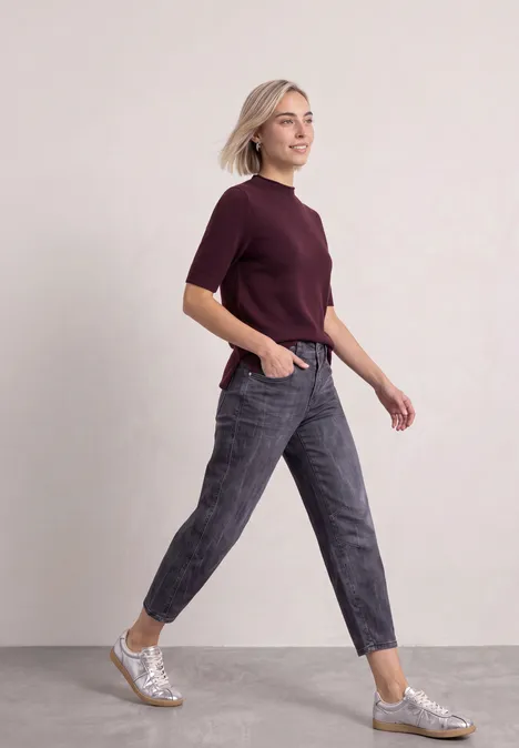 Eine junge Frau in bordeauxfarbenem T-Shirt und grauen Jeans läuft dynamisch, trägt silberne Sneaker.