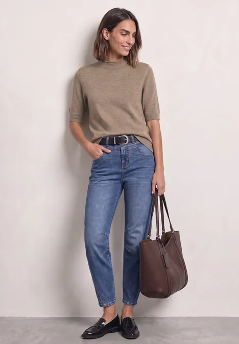 Vrouw in beige trui, blauwe jeans en zwarte loafers met een bruine schoudertas.