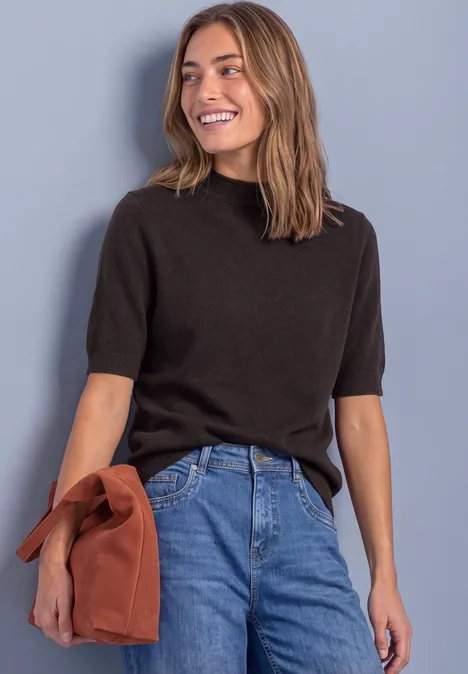 Lachende vrouw in zwarte korte trui en blauwe jeans, met een roestkleurige tas voor een grijze muur.
